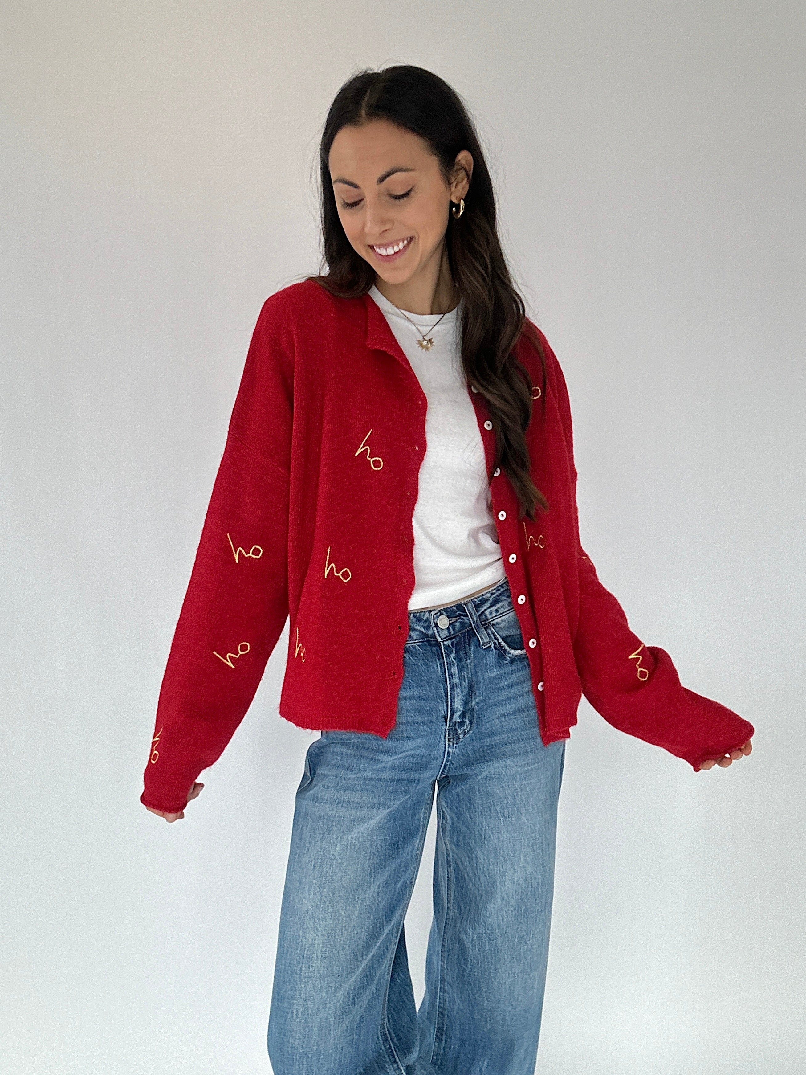 Woman wearing a red cardigan with white buttons and gold embroidered lettering, white shirt, and blue jeans on a plain background