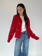 Woman wearing a red cardigan with white buttons and gold embroidered lettering, white shirt, and blue jeans on a plain background