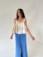 A woman modeling a cream and beige sleeveless top with intricate embroidery and a ruffled peplum hem, paired with wide-leg blue jeans, against a plain off-white studio background.