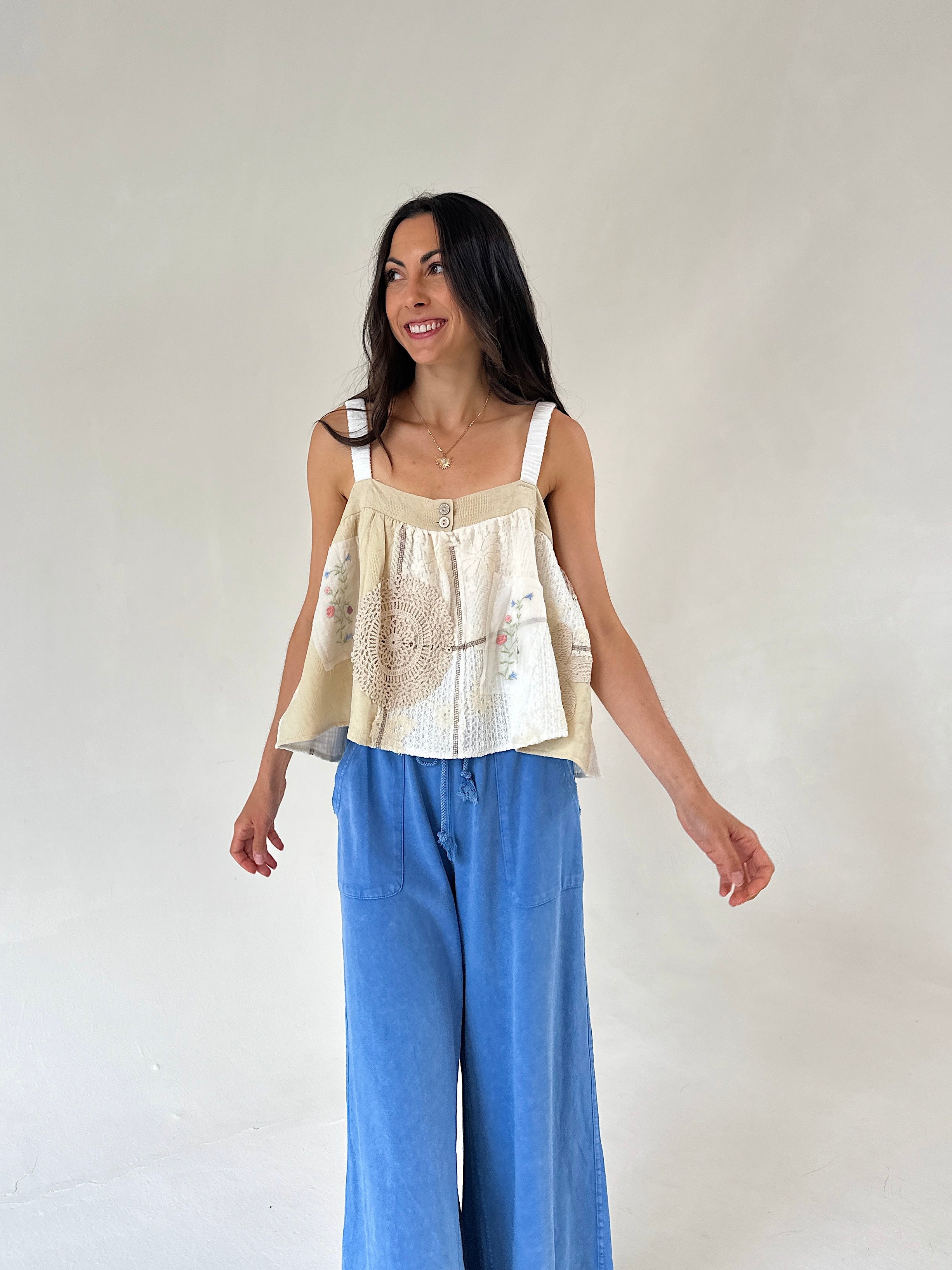A woman modeling a cream and beige sleeveless top with intricate embroidery and a ruffled peplum hem, paired with wide-leg blue jeans, against a plain off-white studio background.