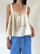 A woman wearing a cream-colored sleeveless top with textured crochet lace panels, floral embroidery, and white shoulder straps, paired with blue drawstring pants against a plain light background.