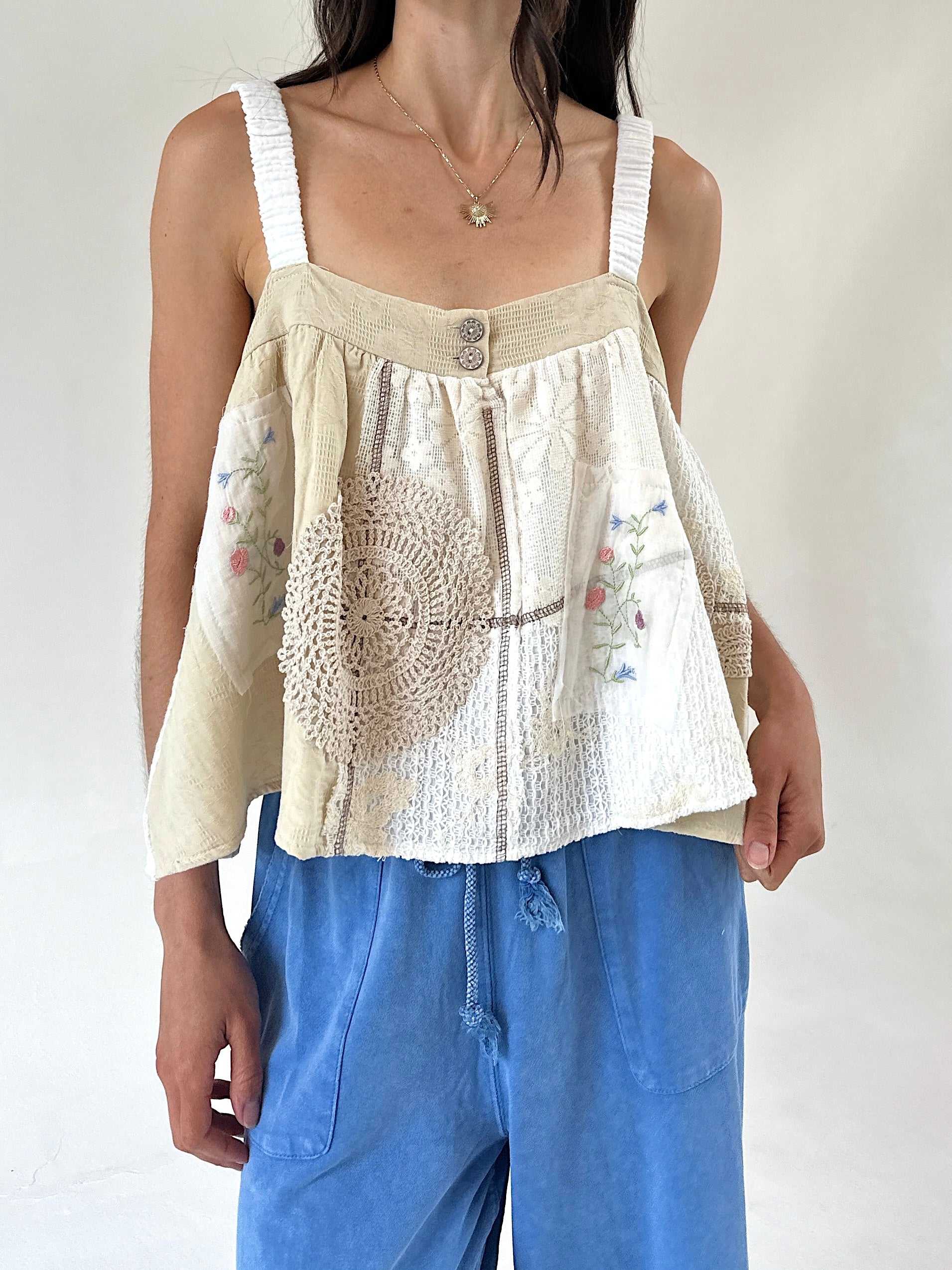 A woman wearing a cream-colored sleeveless top with textured crochet lace panels, floral embroidery, and white shoulder straps, paired with blue drawstring pants against a plain light background.