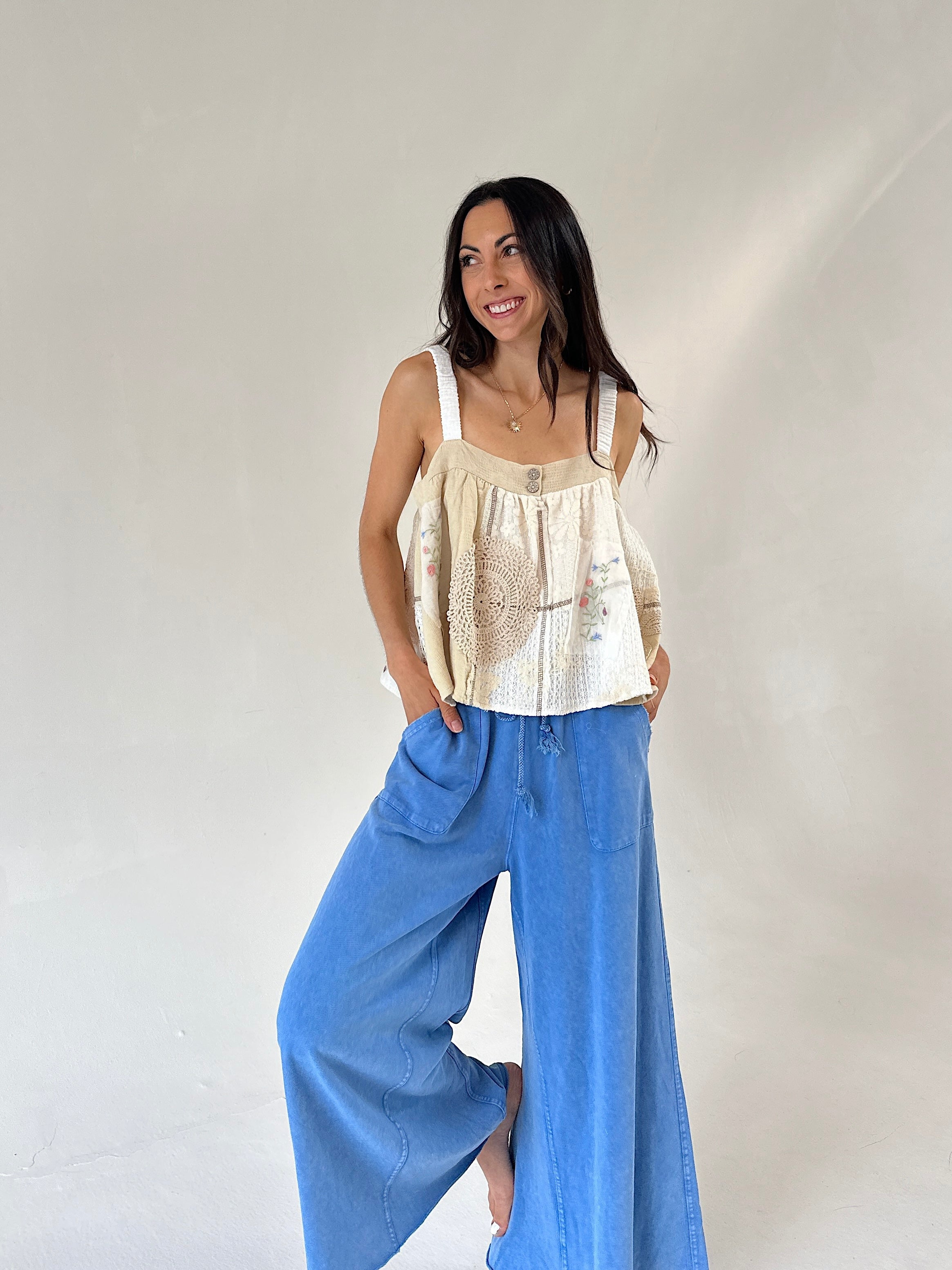 A woman modeling a cream sleeveless top with crochet detailing and wide-leg blue jeans against a plain light gray studio background.