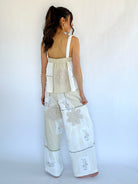 Woman wearing a beige sleeveless top and white pants with floral patterns against a white background