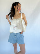 Woman wearing a sleeveless top with a floral pattern and denim shorts against a plain background