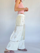 Person wearing a pink top and white pants with floral embroidery on a plain background from side