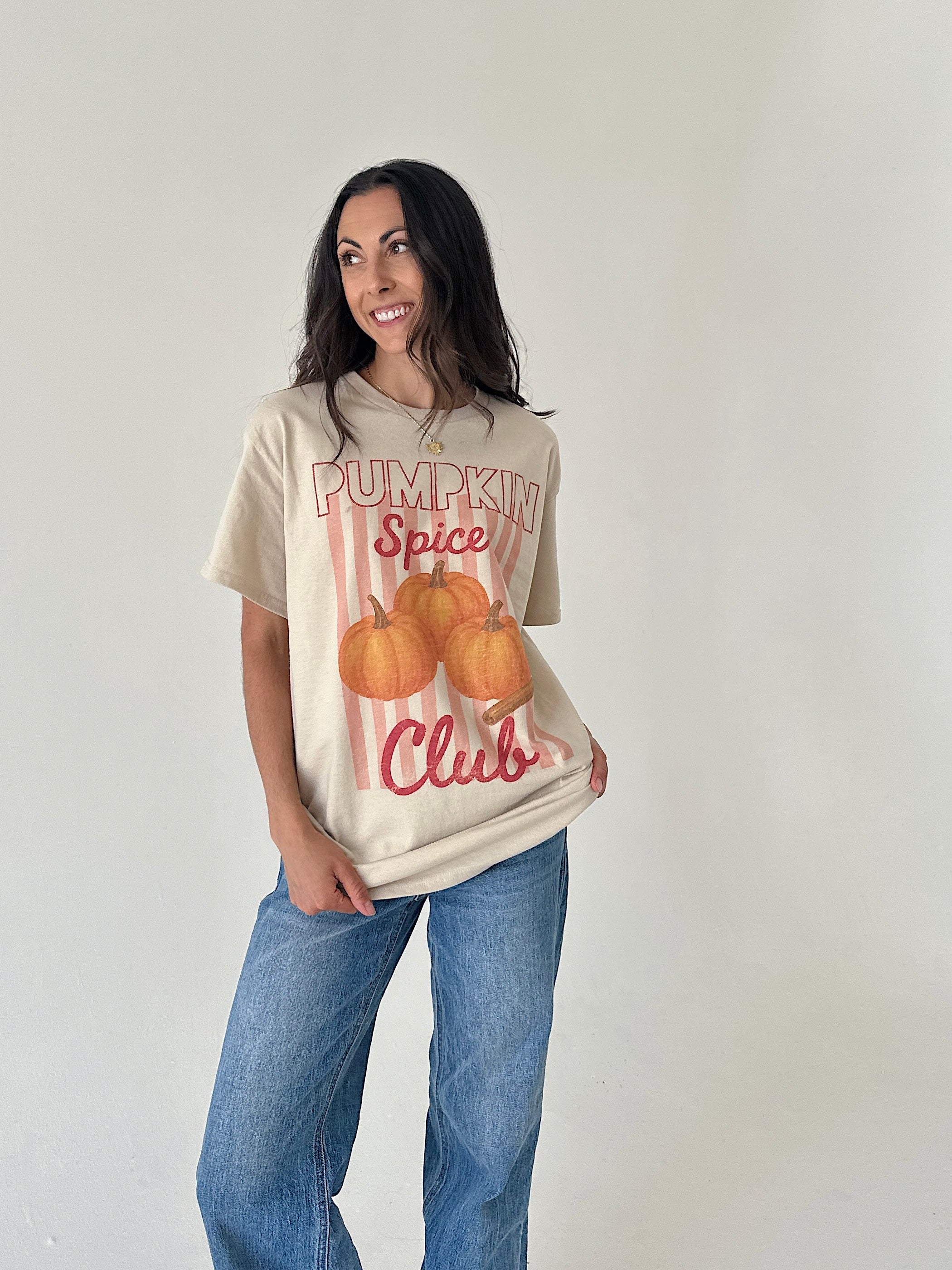 A woman modeling a beige graphic t-shirt with 'Pumpkin Spice Club' and pumpkins printed on the front, paired with blue jeans, against a plain off-white background.