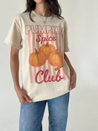 A woman wearing a cream-colored graphic t-shirt with 'Pumpkin Spice Club' text and an illustration of pumpkins, paired with blue jeans, against a plain light background.