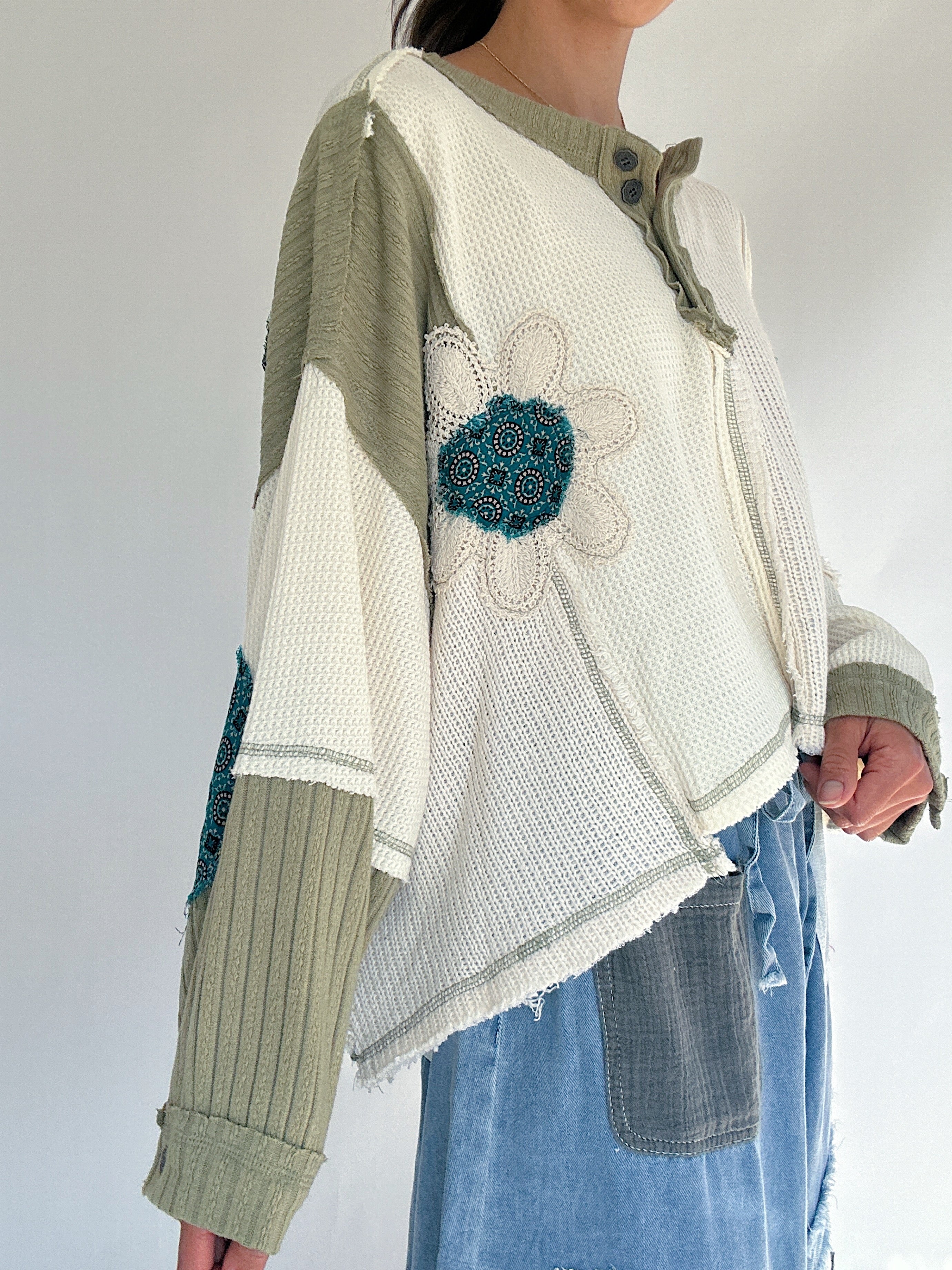 Person wearing a patchwork knit sweater with a floral patch on a plain background showing a side view