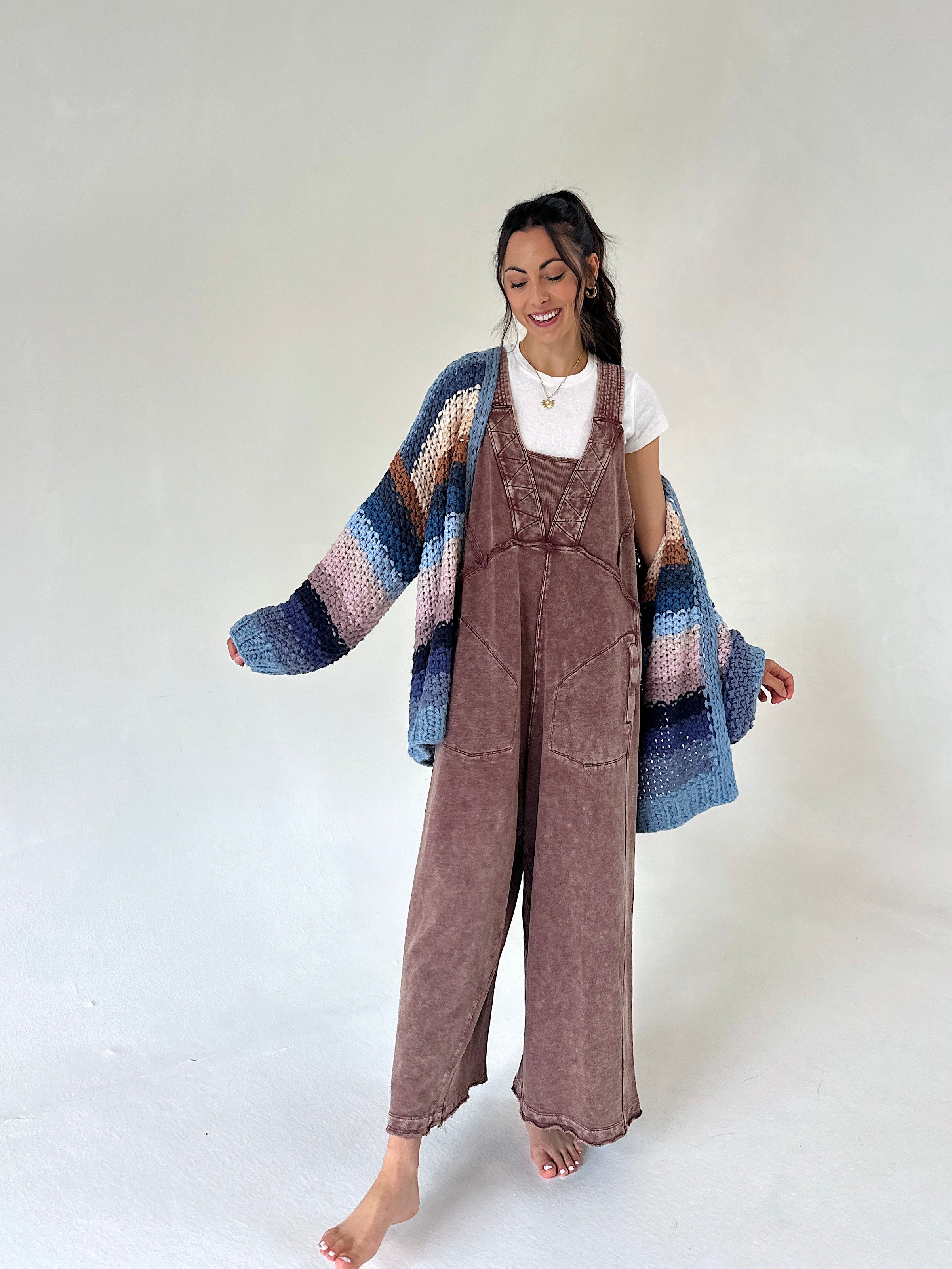 A woman stands barefoot against a plain light background wearing a brown terry knit overall jumpsuit, a white t-shirt, and a chunky knit cardigan with blue, pink, and purple stripes.
