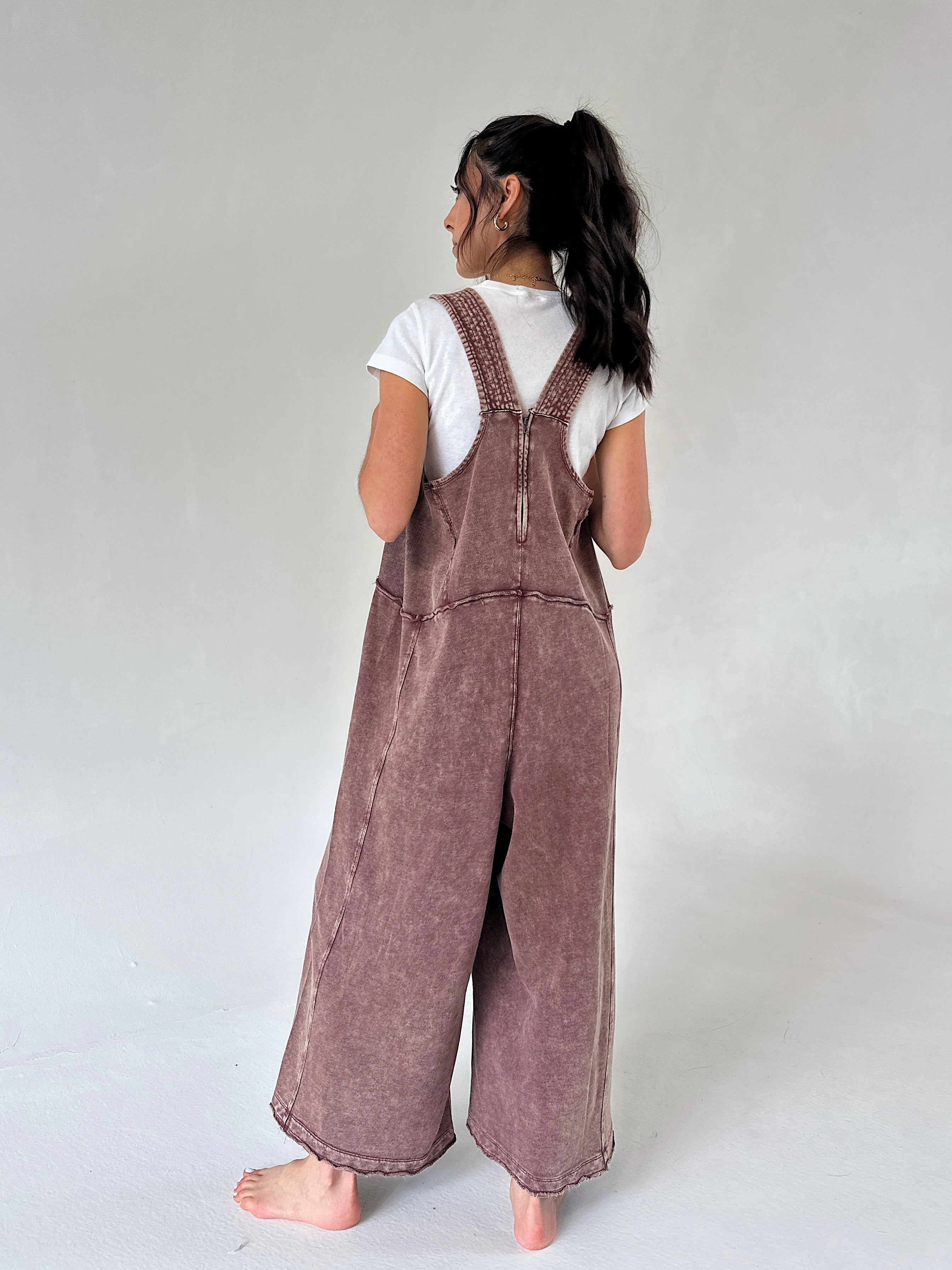A model shown from the back wearing a dusty mauve-brown wide-leg terry overall jumpsuit over a white t-shirt, standing barefoot against a neutral light gray studio background.