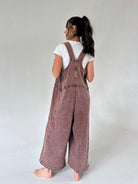 A model shown from the back wearing a dusty mauve-brown wide-leg terry overall jumpsuit over a white t-shirt, standing barefoot against a neutral light gray studio background.