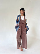 A woman stands barefoot against a plain light gray studio background, wearing a brown terry overall jumpsuit with a white top and a chunky knit cardigan in blue, beige, and brown tones.