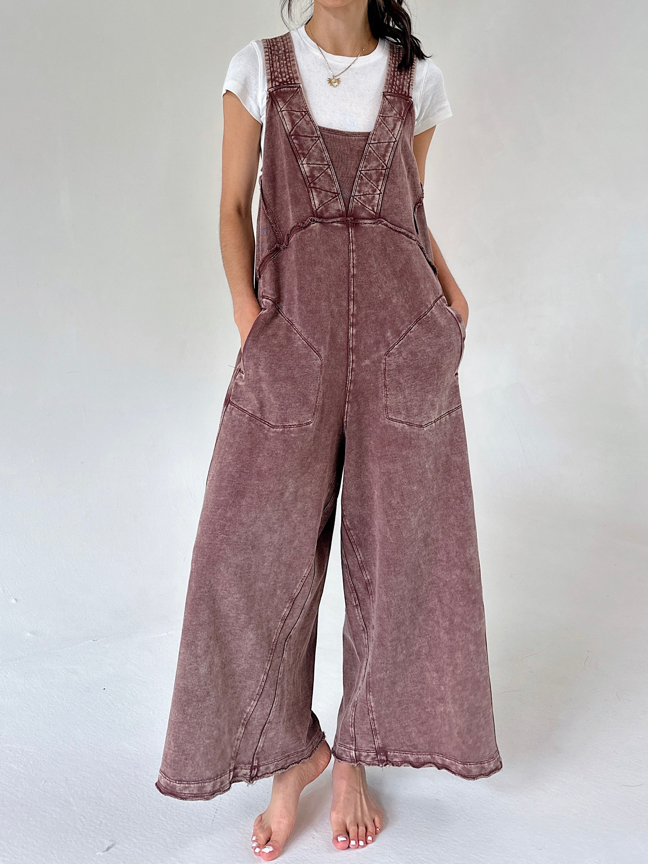 A woman wearing a brownish mauve wide-leg terry overall jumpsuit over a white t-shirt, standing against a light gray studio background.
