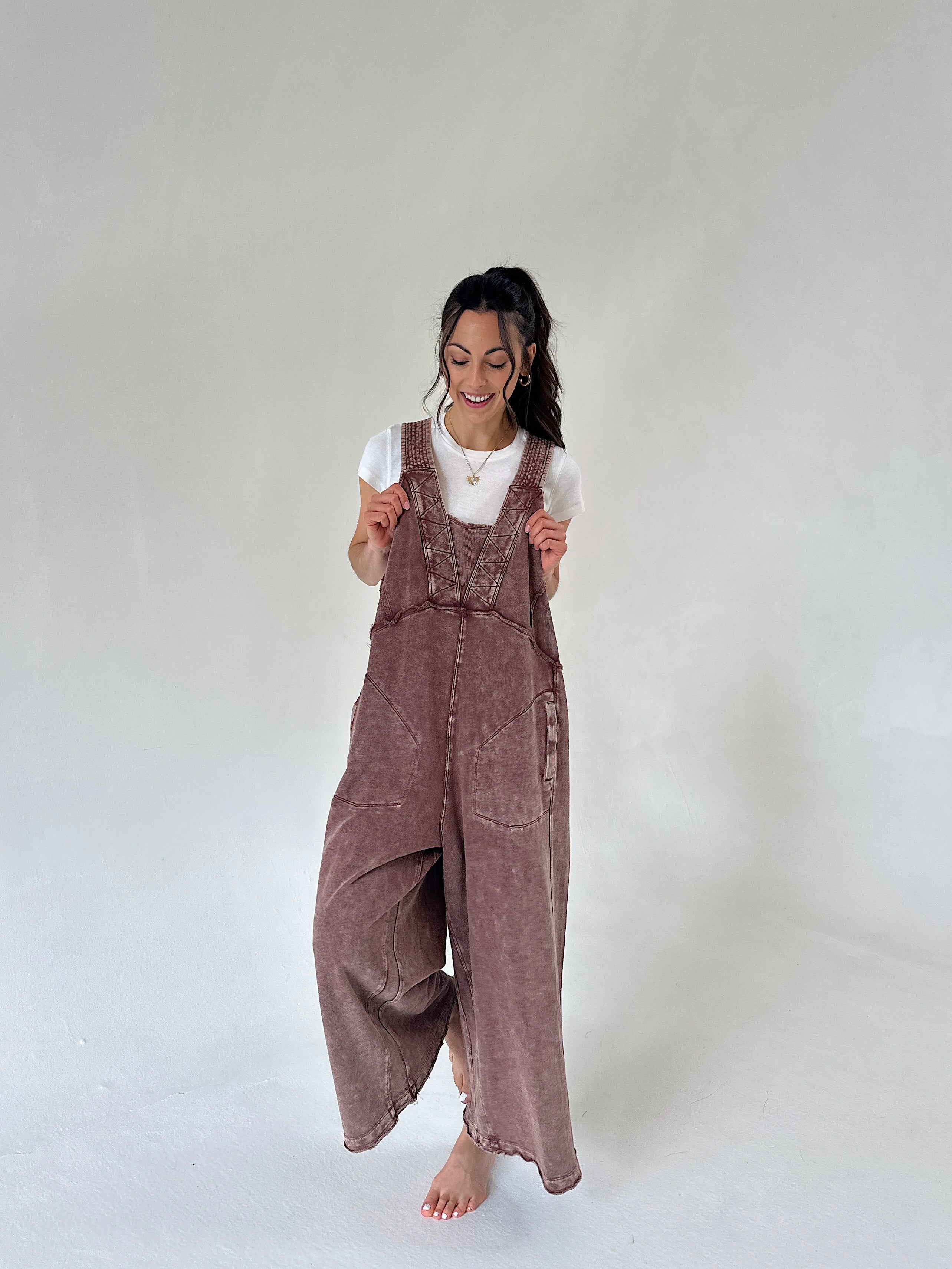 A woman wearing a dusty mauve-brown terry knit overall jumpsuit over a white t-shirt, standing barefoot against a neutral light gray studio background.