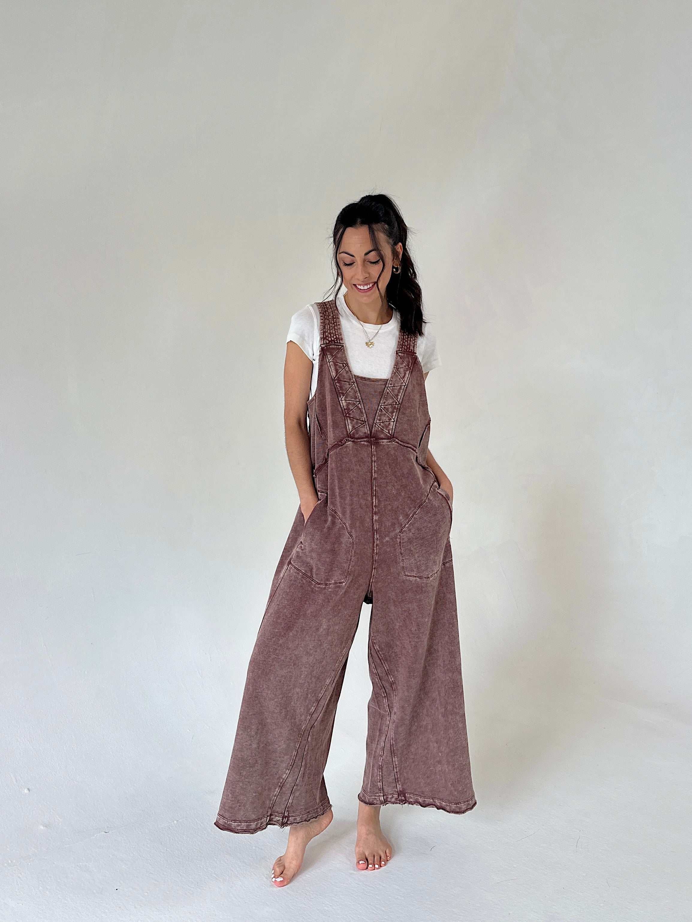 A woman modeling a brownish mauve wide-leg terry jumpsuit over a white t-shirt, standing against a neutral light gray studio background.