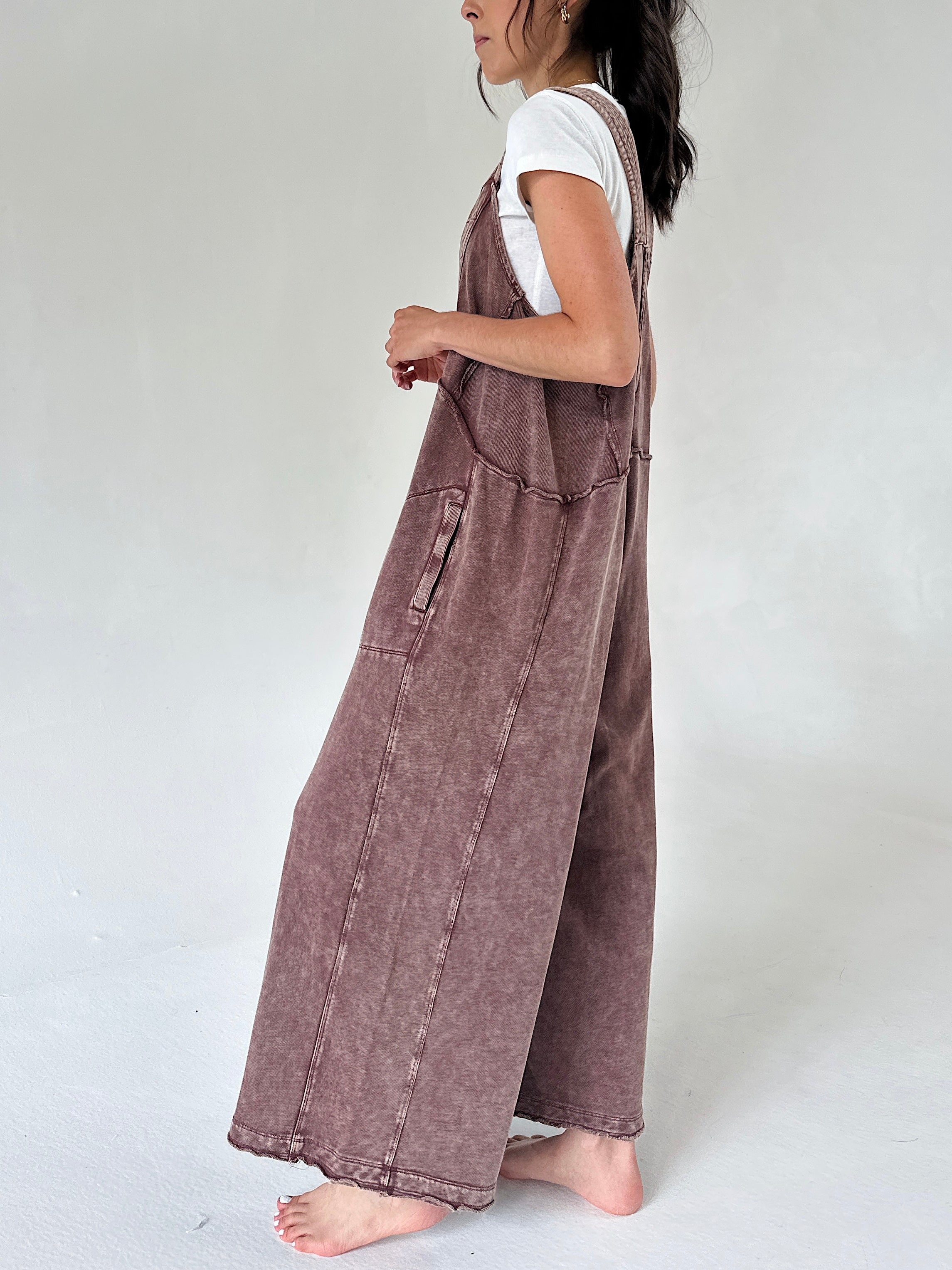 A woman shown from the side wearing a dusty brownish mauve wide-leg terry knit overall jumpsuit over a white t-shirt, standing barefoot against a plain light gray studio background.