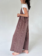 A woman shown from the side wearing a dusty brownish mauve wide-leg terry knit overall jumpsuit over a white t-shirt, standing barefoot against a plain light gray studio background.