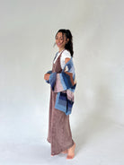 A woman stands barefoot against a plain light gray studio background, wearing a long mauve-brown jumpsuit over a white top and holding a chunky knit patchwork throw in blue, pink, and brown tones.