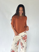 A woman modeling a short-sleeve knit top in a warm rust orange color, paired with cream wide-leg pants featuring a red and gray floral pattern, against a plain white studio background.
