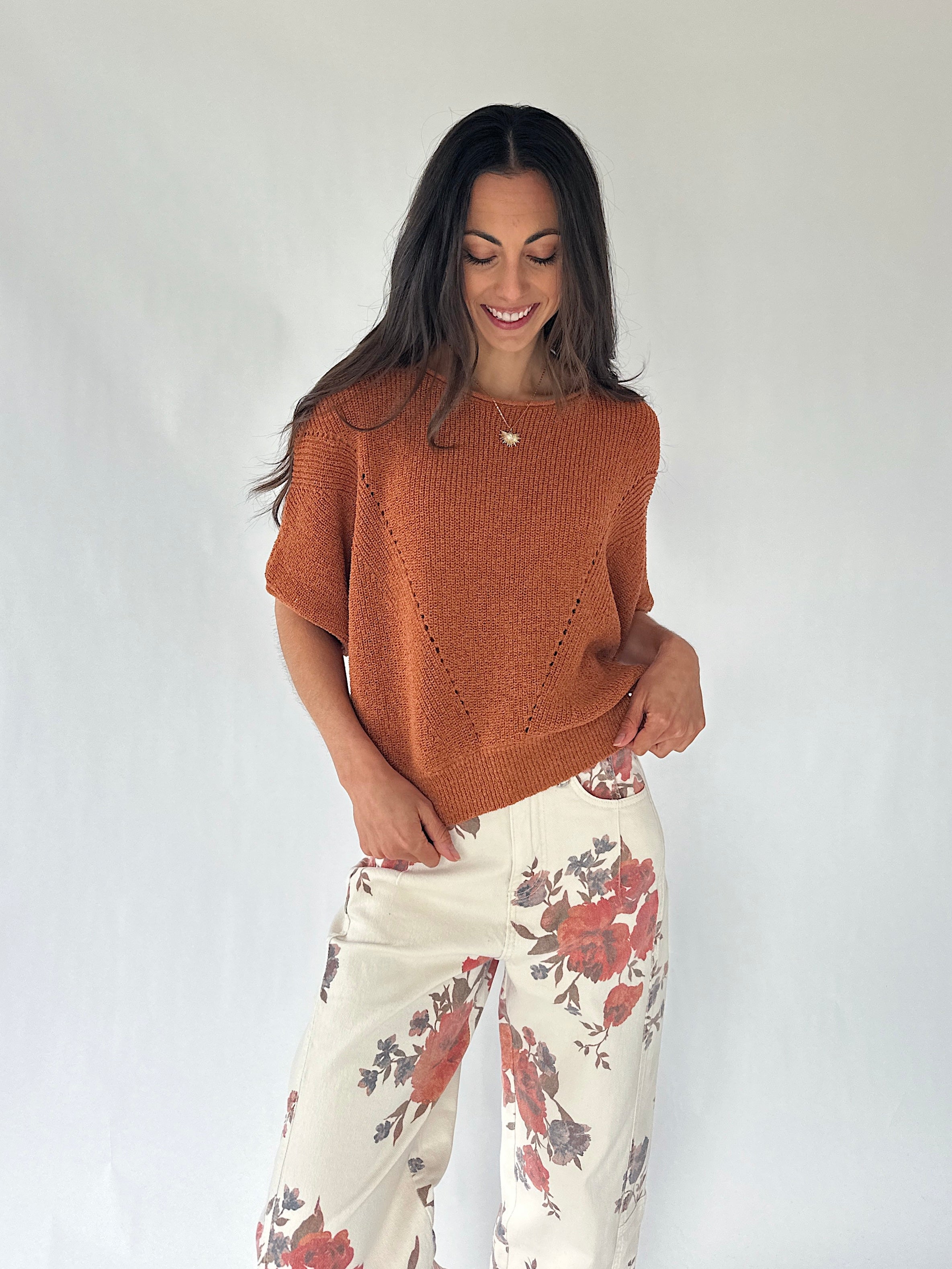 A woman modeling a short-sleeve knit top in a warm rust orange color, paired with cream wide-leg pants featuring a red and gray floral pattern, against a plain white studio background.