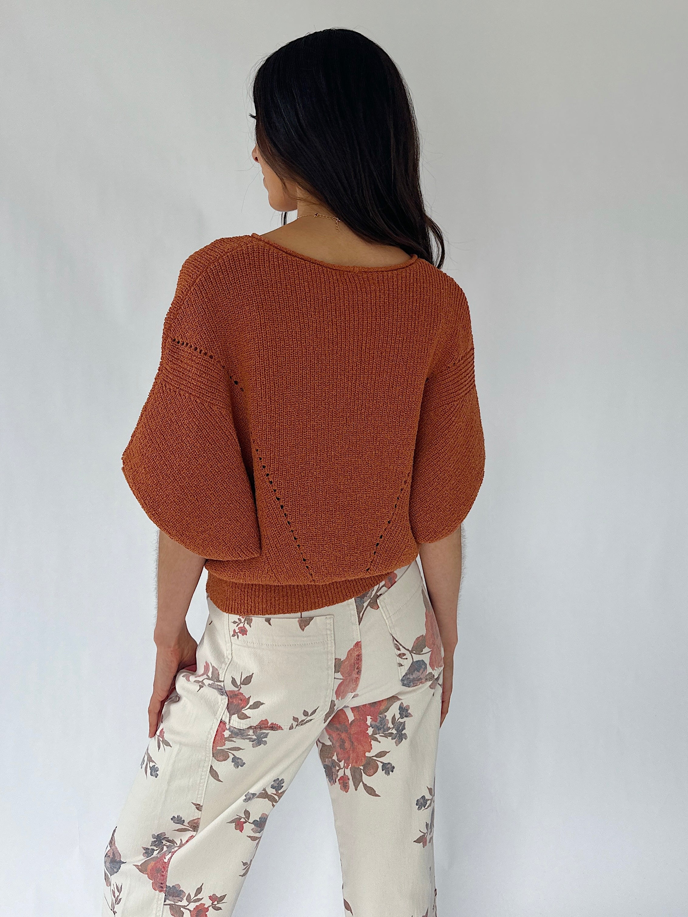 A woman shown from behind wearing a rust-orange knit short-sleeve top and cream floral pants against a plain light gray background.