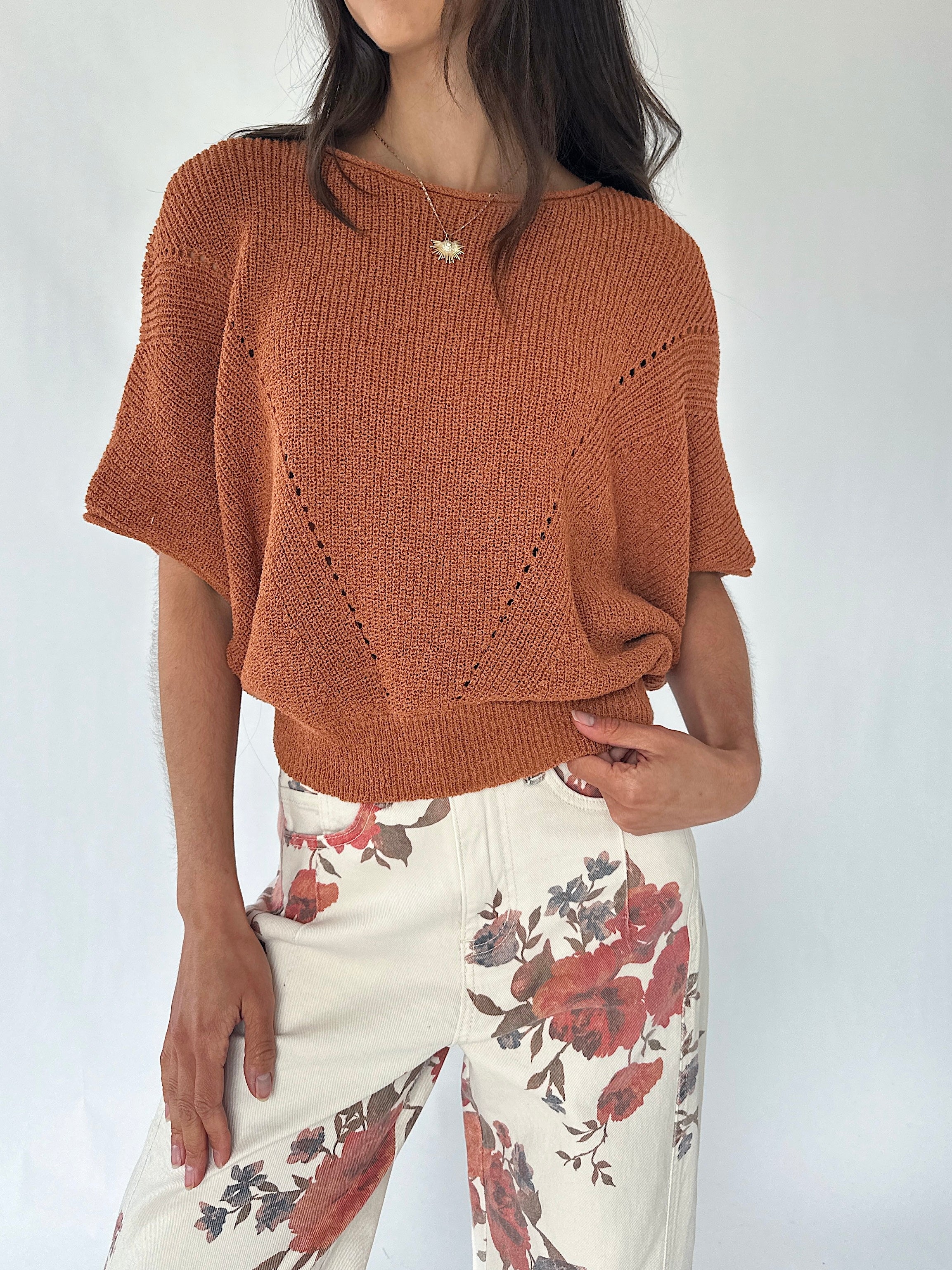 A woman wearing a rust-orange knit short-sleeve sweater with textured openwork details and cream floral print pants, photographed against a plain light background.