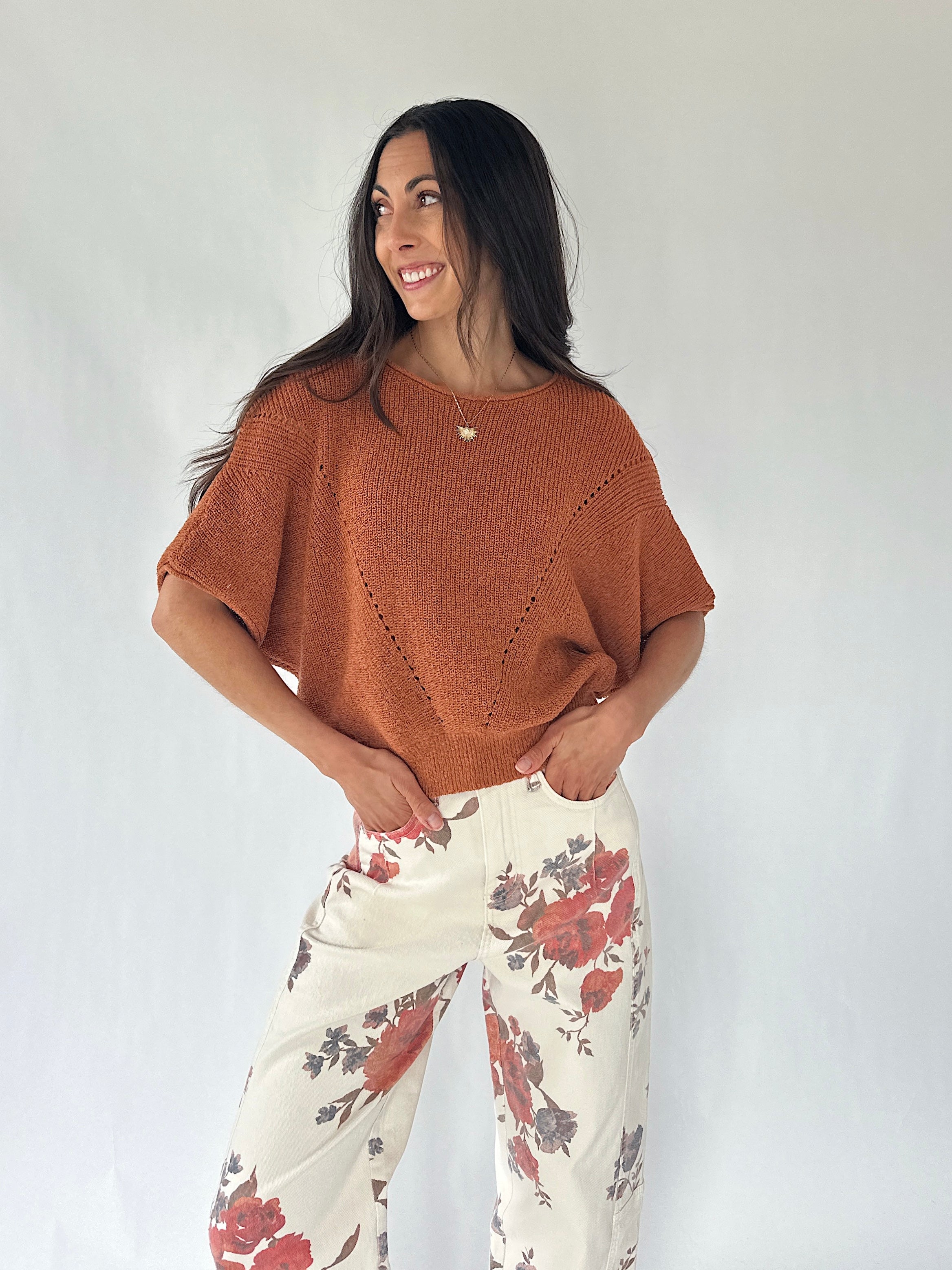 A woman modeling a short-sleeve knit top in a burnt orange color, paired with cream-colored pants featuring a red and gray floral pattern, set against a plain light gray background.