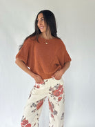A woman modeling a short-sleeve knit top in a burnt orange color, paired with cream-colored pants featuring a red and gray floral pattern, set against a plain light gray background.