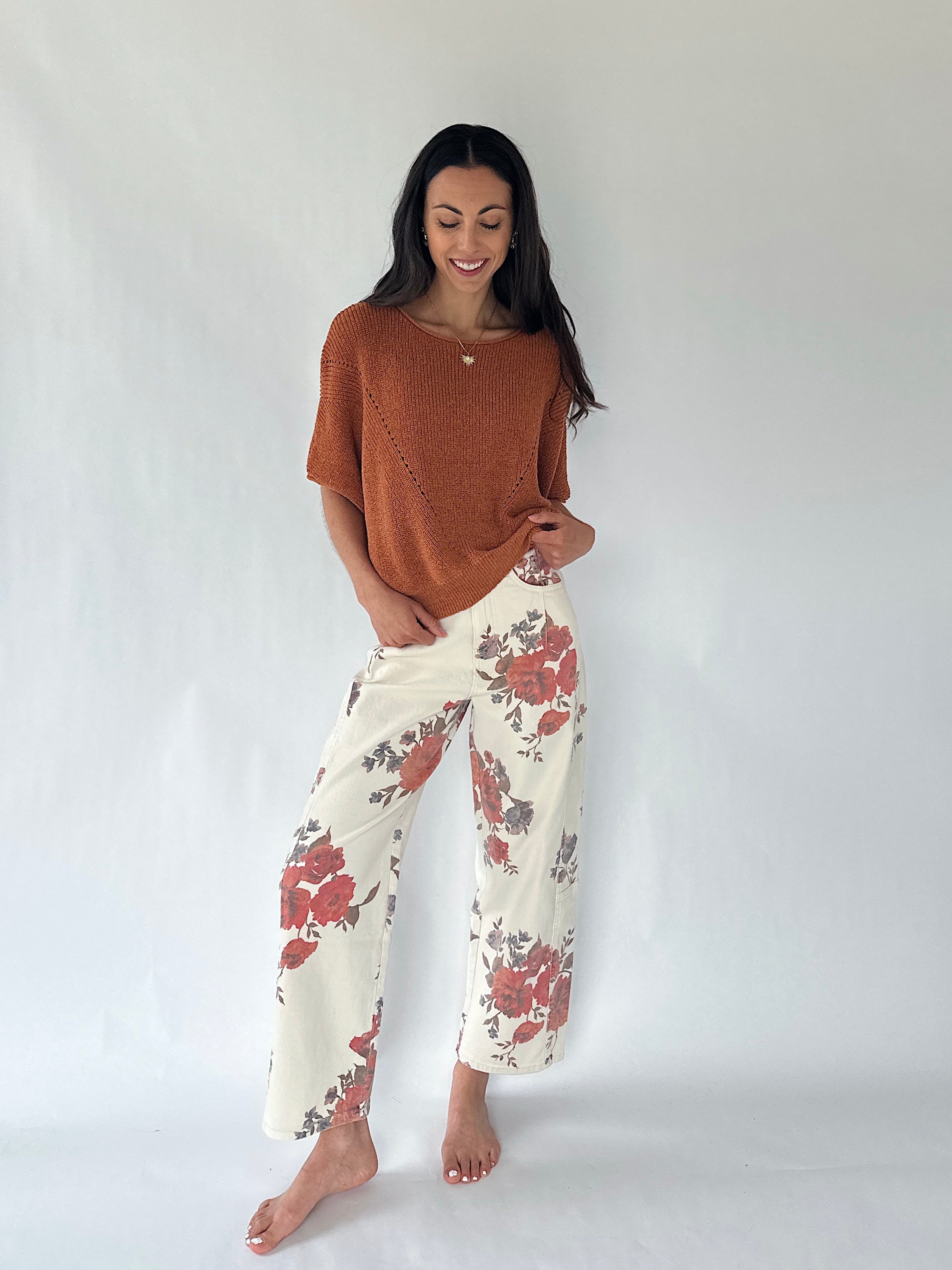 A woman stands barefoot against a plain light gray studio background, wearing a rust-orange short-sleeve knit top and cream wide-leg pants with a red and gray floral print.