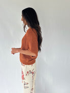 A woman shown in side profile wearing a textured rust-orange knit sweater and cream floral pants against a plain light gray wall.
