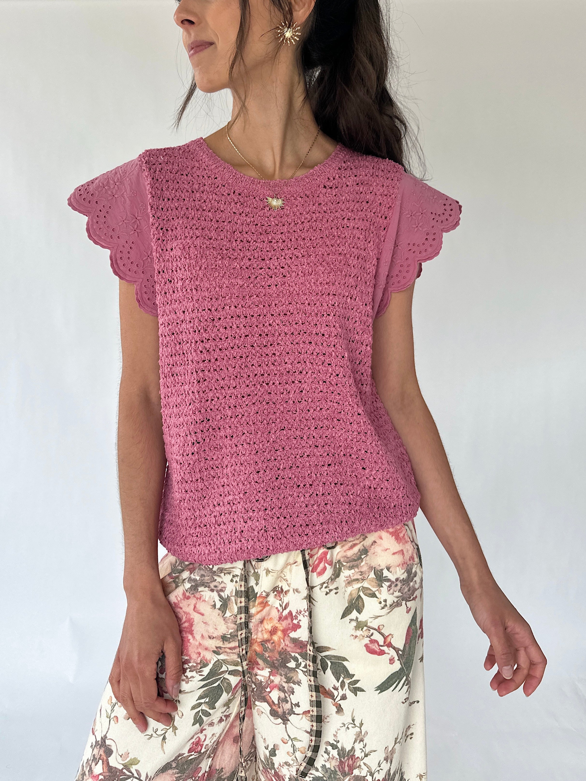 A woman wearing a pink knit short-sleeve top with scalloped edges and floral print pants, photographed against a plain light background.