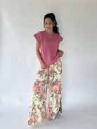A woman modeling a pink short-sleeve textured top and wide-leg floral pants with a pink, red, and green botanical print on a cream background, standing against a white studio backdrop.