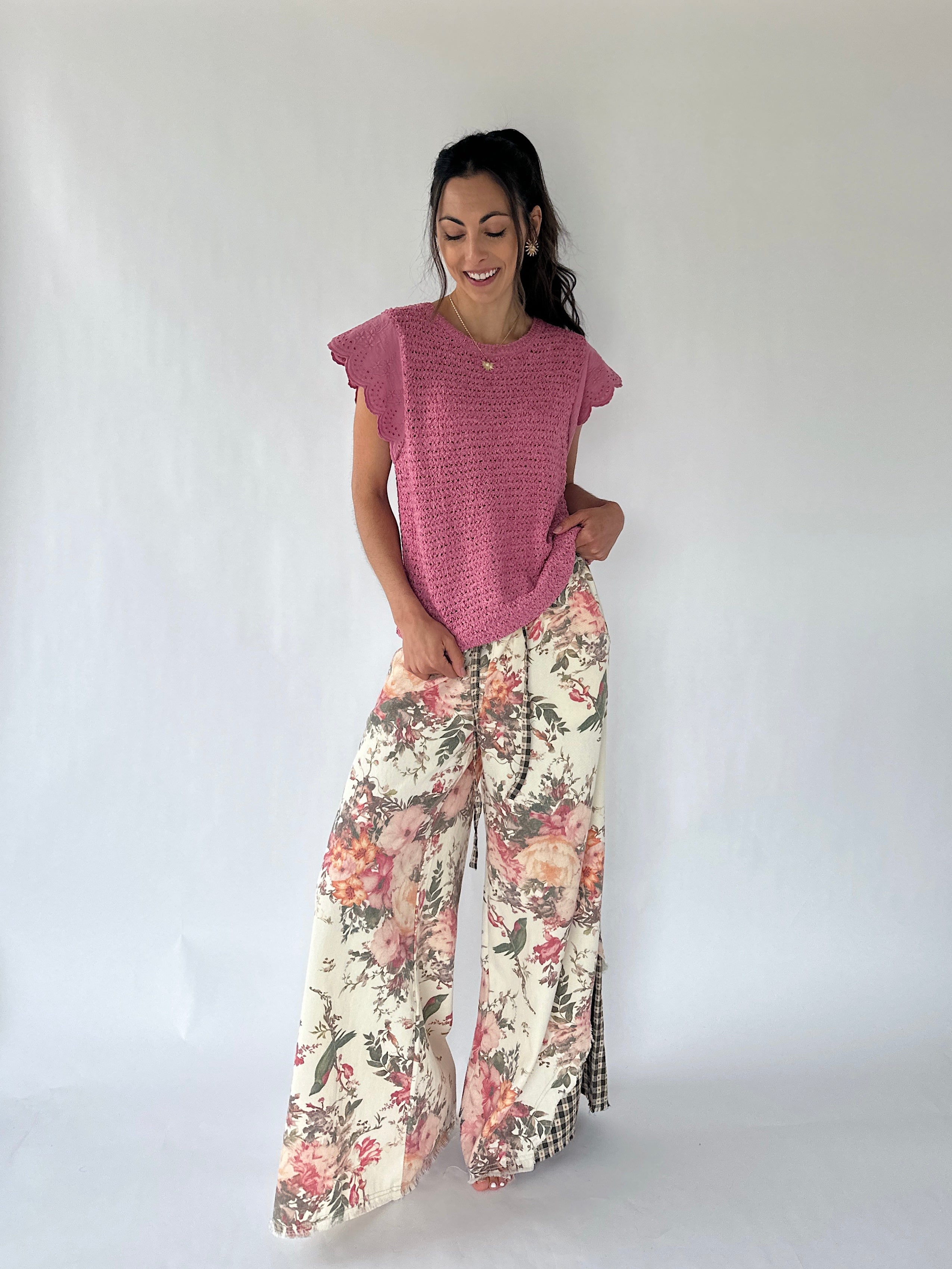 A woman modeling a pink short-sleeve textured top and wide-leg floral pants with a pink, red, and green botanical print on a cream background, standing against a white studio backdrop.