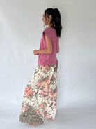 A woman stands in profile wearing a pink sleeveless knit top and floral pants with a gingham hem, photographed against a plain light gray studio background.