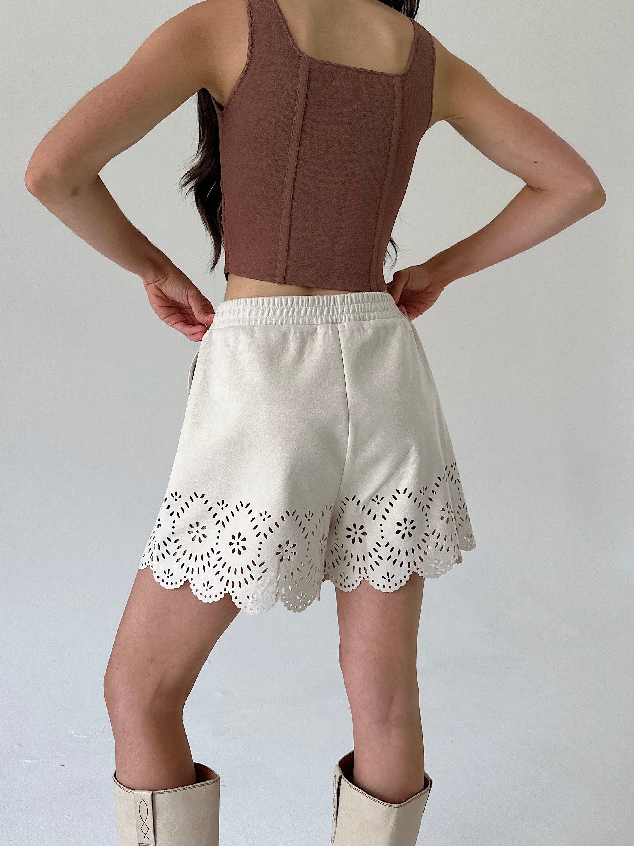 A model shown from the back wearing a brown sleeveless crop top and cream-colored shorts with a scalloped eyelet lace hem, paired with cream boots against a plain light gray background.