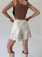 A model shown from the back wearing a brown sleeveless crop top and cream-colored shorts with a scalloped eyelet lace hem, paired with cream boots against a plain light gray background.