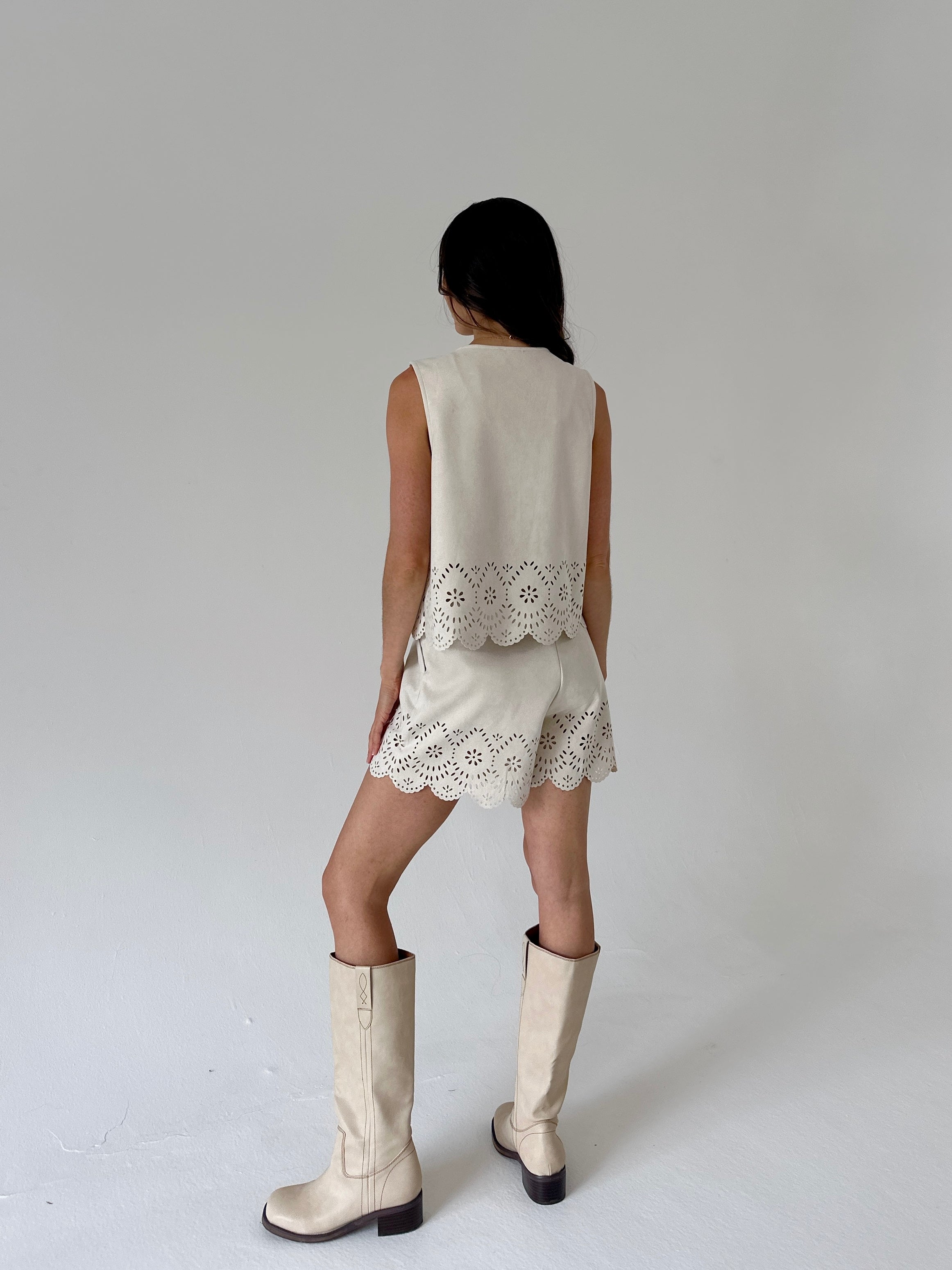 A woman is shown from behind wearing a sleeveless cream suede vest and matching shorts with scalloped eyelet lace trim, paired with cream-colored knee-high boots, against a neutral light gray studio background.