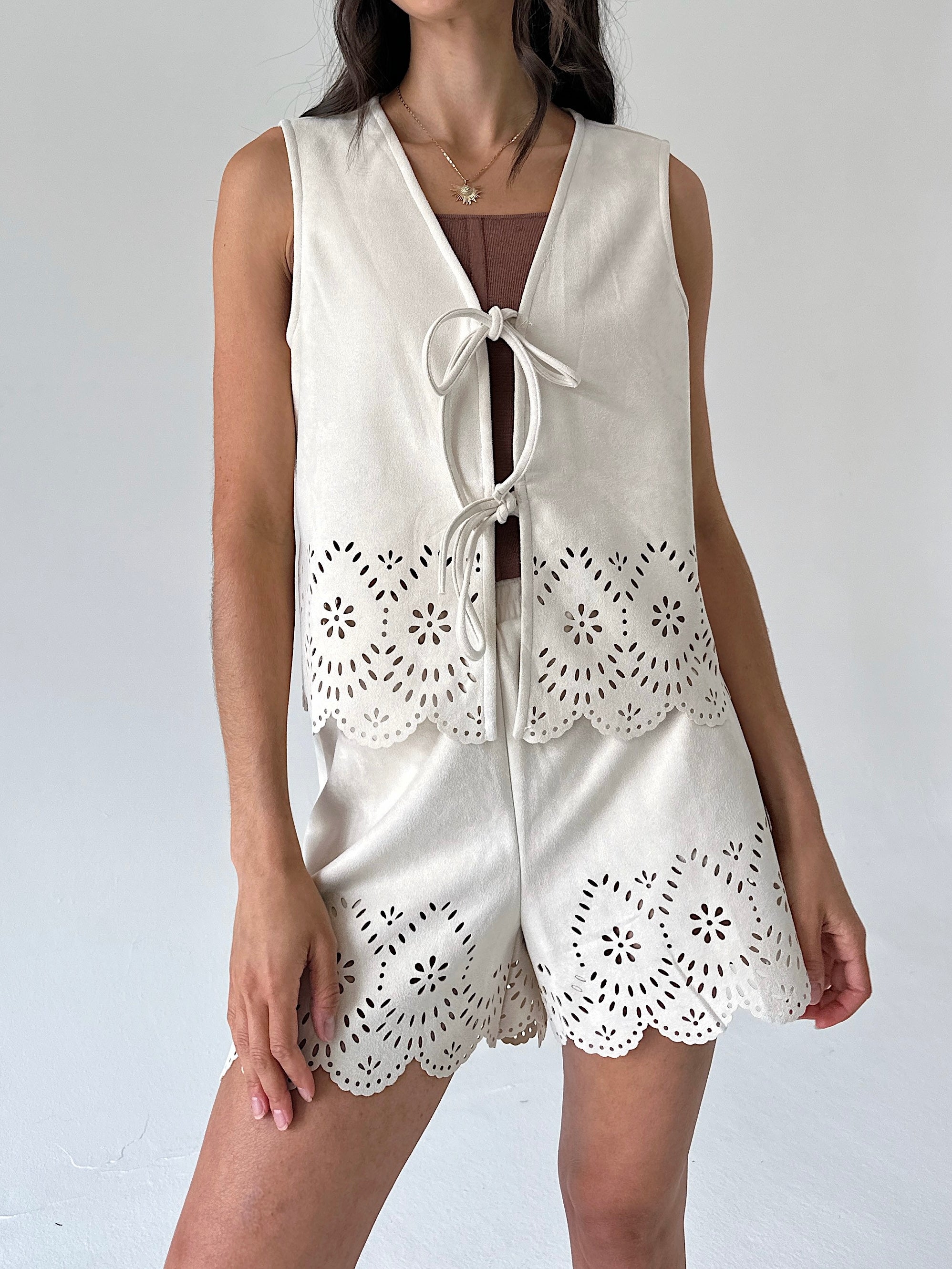 A woman wearing a sleeveless cream-colored vest with intricate eyelet lace detailing and a front tie closure, layered over a brown top with matching shorts