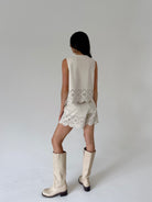 A woman is shown from behind wearing a sleeveless cream suede vest and matching shorts with scalloped eyelet lace trim, paired with cream-colored knee-high boots, against a neutral light gray studio background.