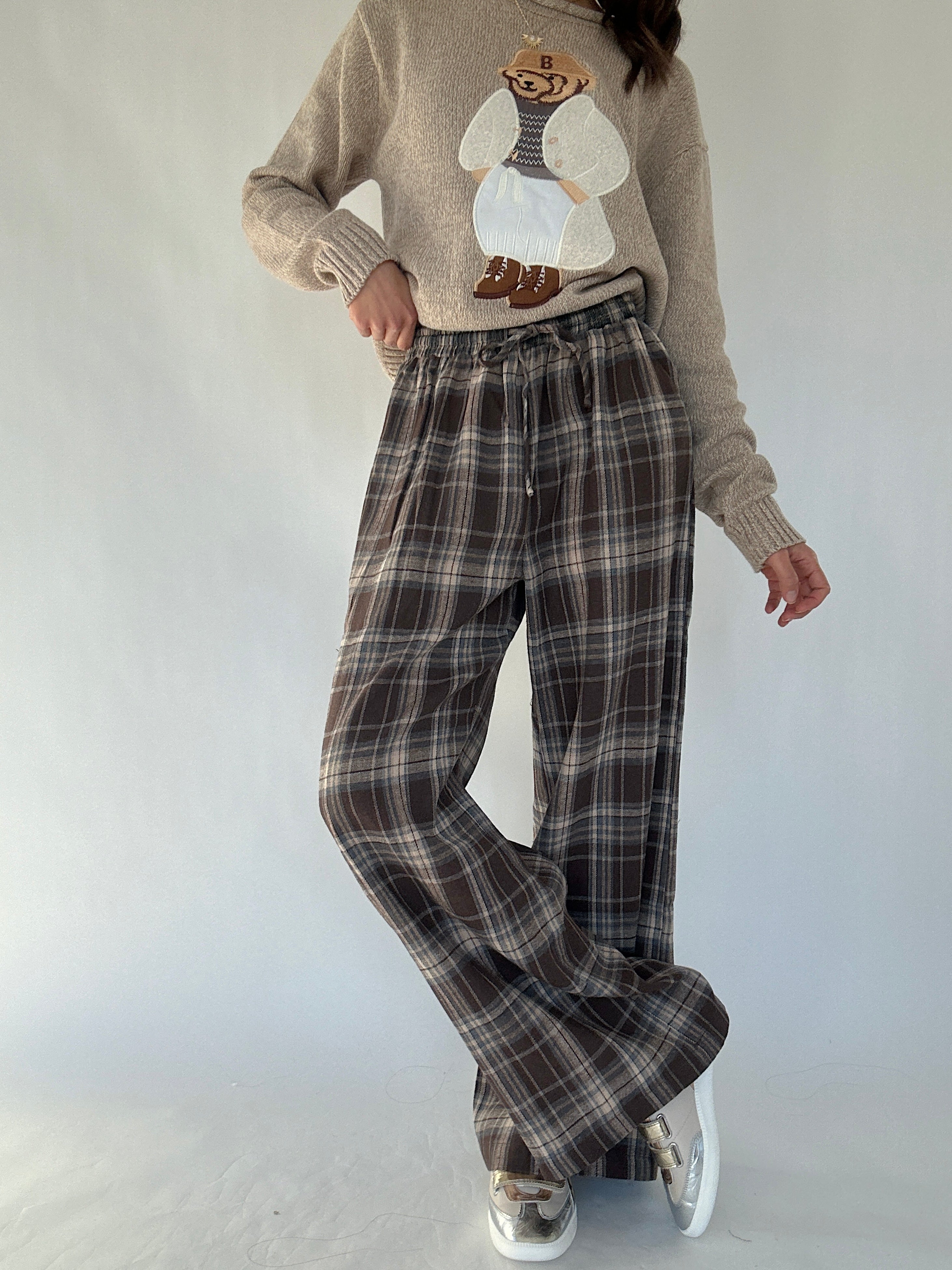Person wearing a beige sweater with a teddy bear design and brown plaid pants with crossed legs on a white background