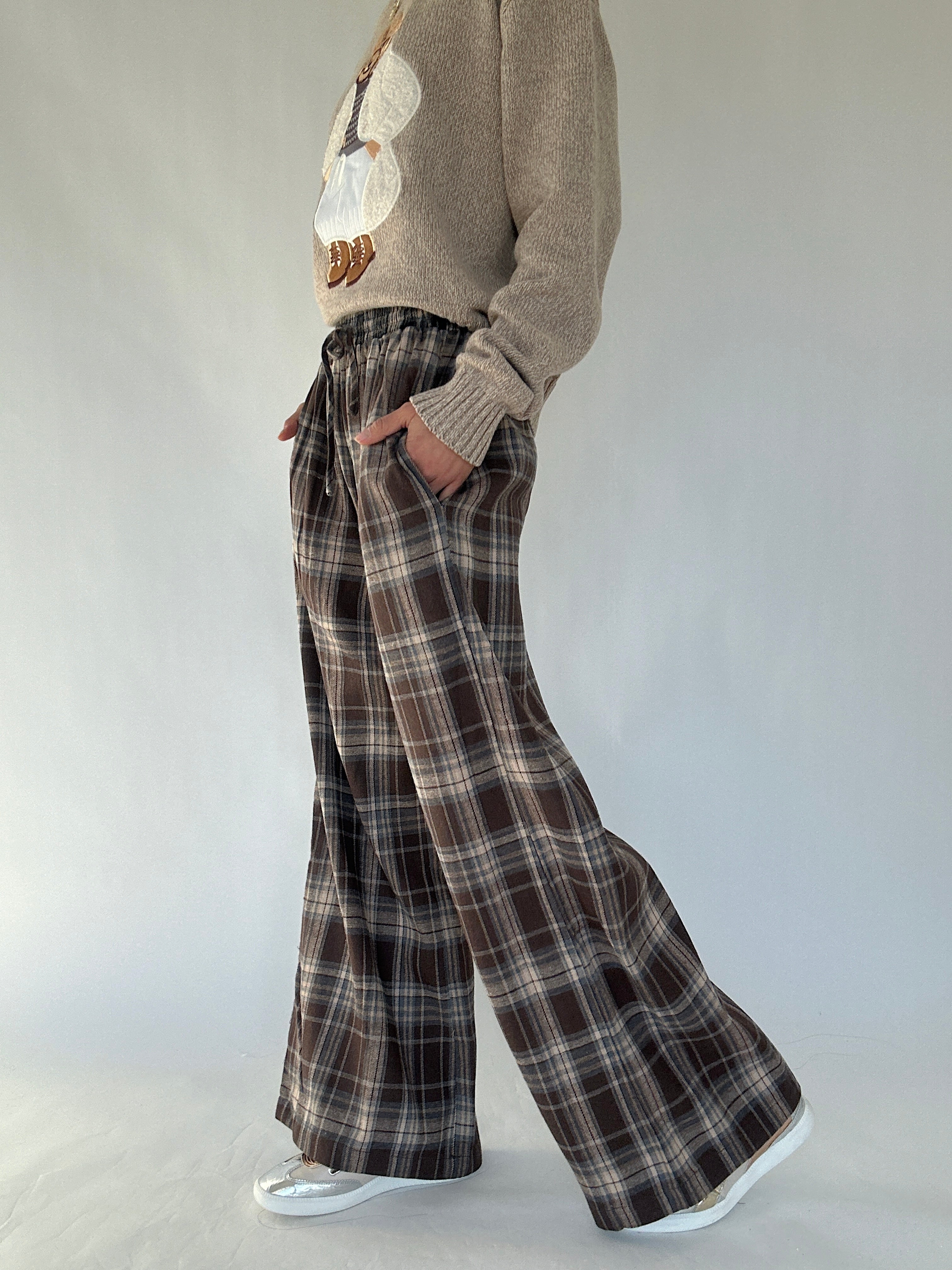 Person wearing a brown sweater and brown plaid pants with hands in side pockets on a plain background