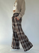 Person wearing a brown sweater and brown plaid pants with hands in side pockets on a plain background