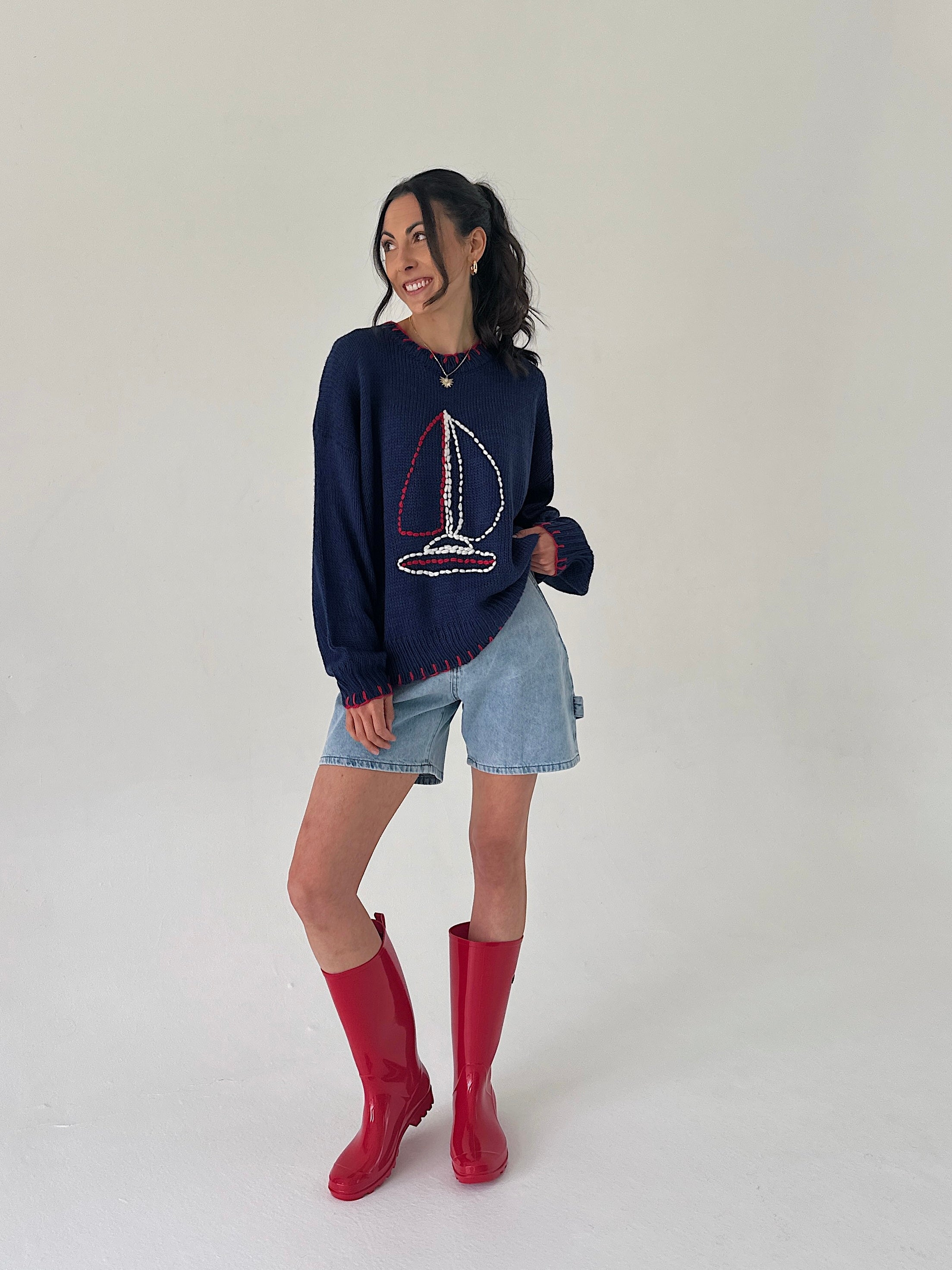 Woman wearing a navy sweater with a sailboat design, denim shorts, and red rain boots on a white background