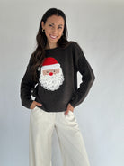 Woman wearing a dark brown long sleeve sweater with a Santa Claus face embroidered design and cream pants on a plain background