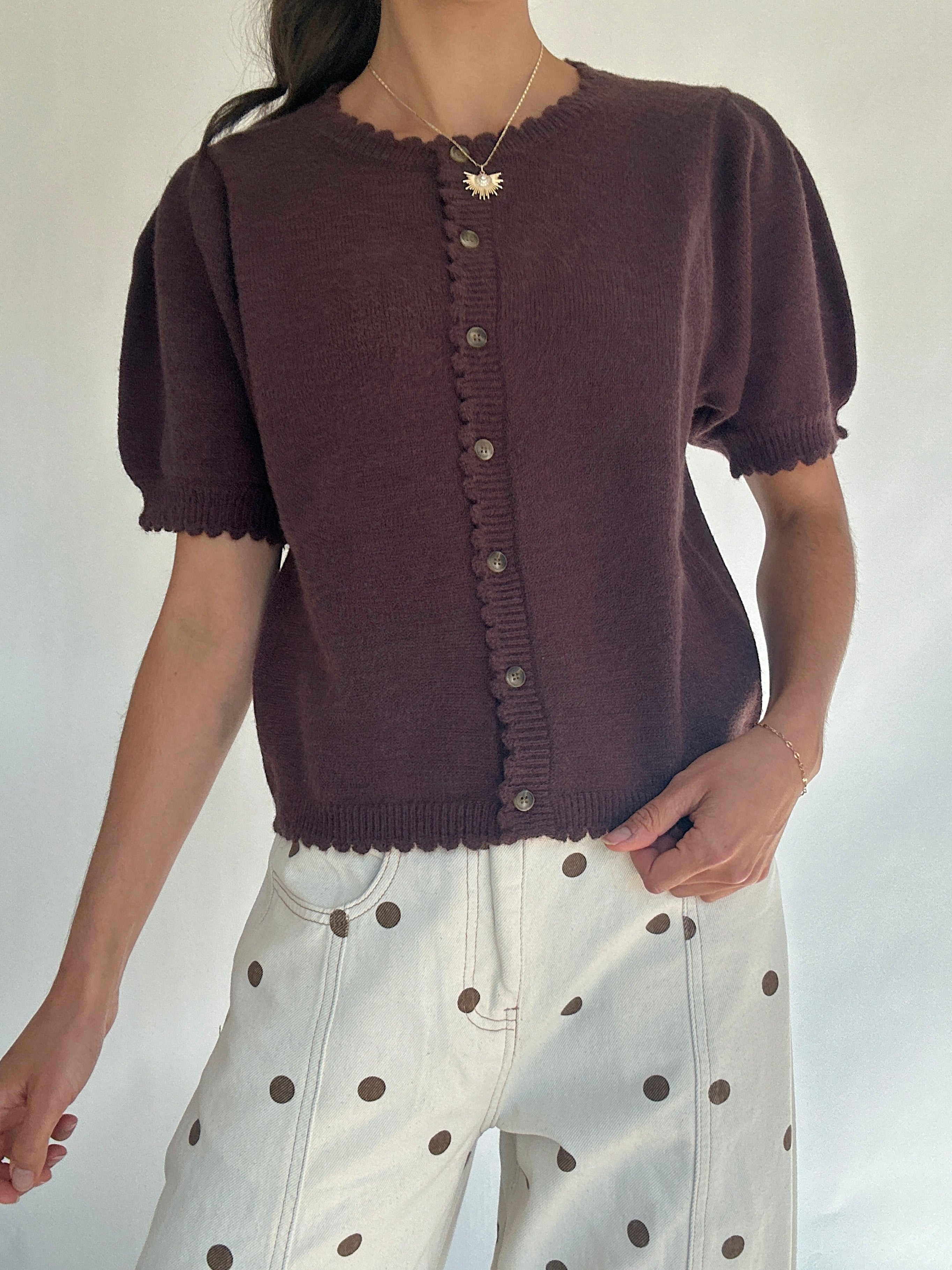 Person wearing a brown knitted short sleeve cardigan with white pants featuring brown dots on a plain background