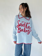 Woman wearing light blue collared sweater with red "Santa Baby" lettering and jeans on a white background