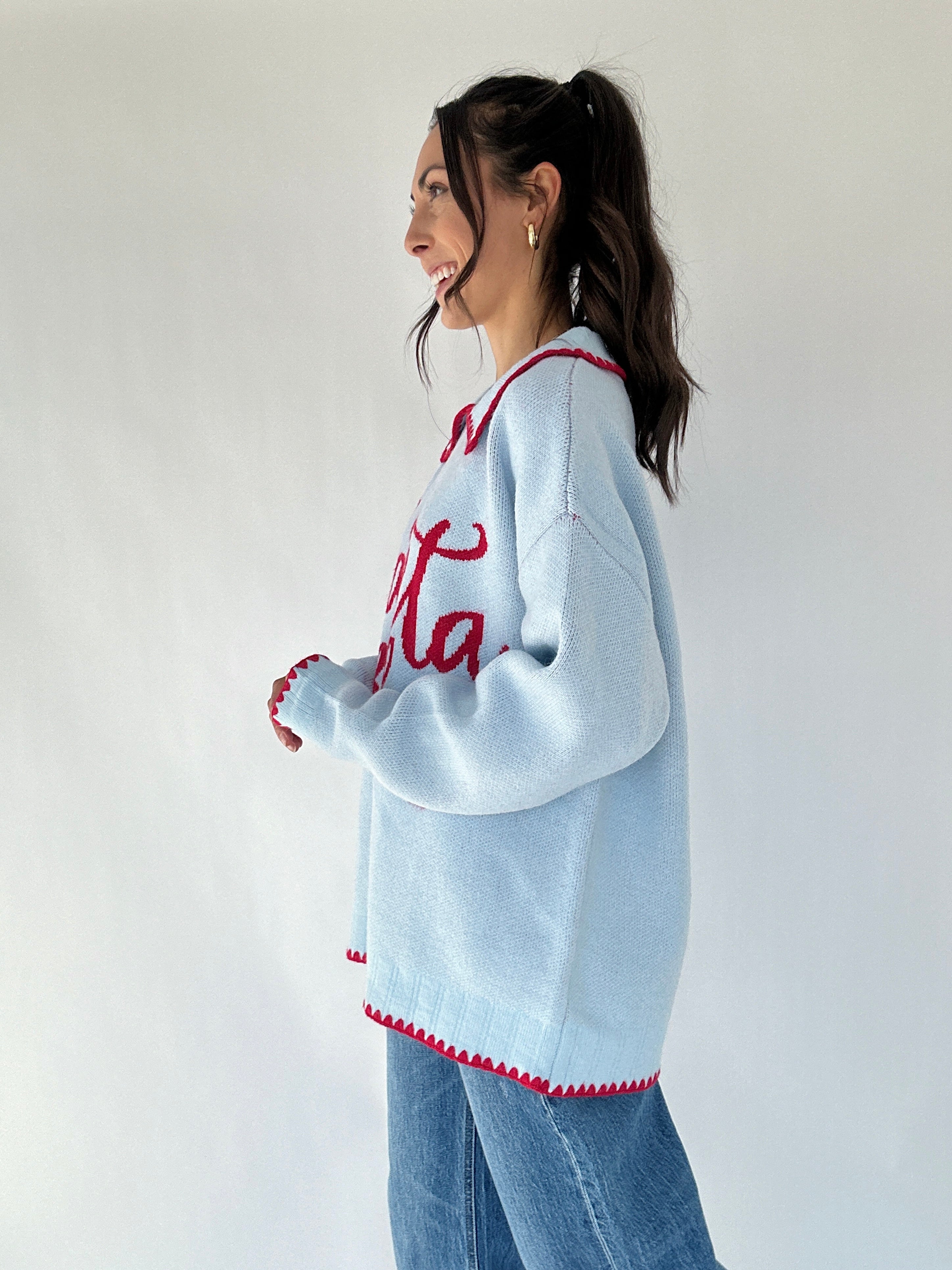 Woman wearing a light blue sweater with red text and white trim, standing against a plain background. side view
