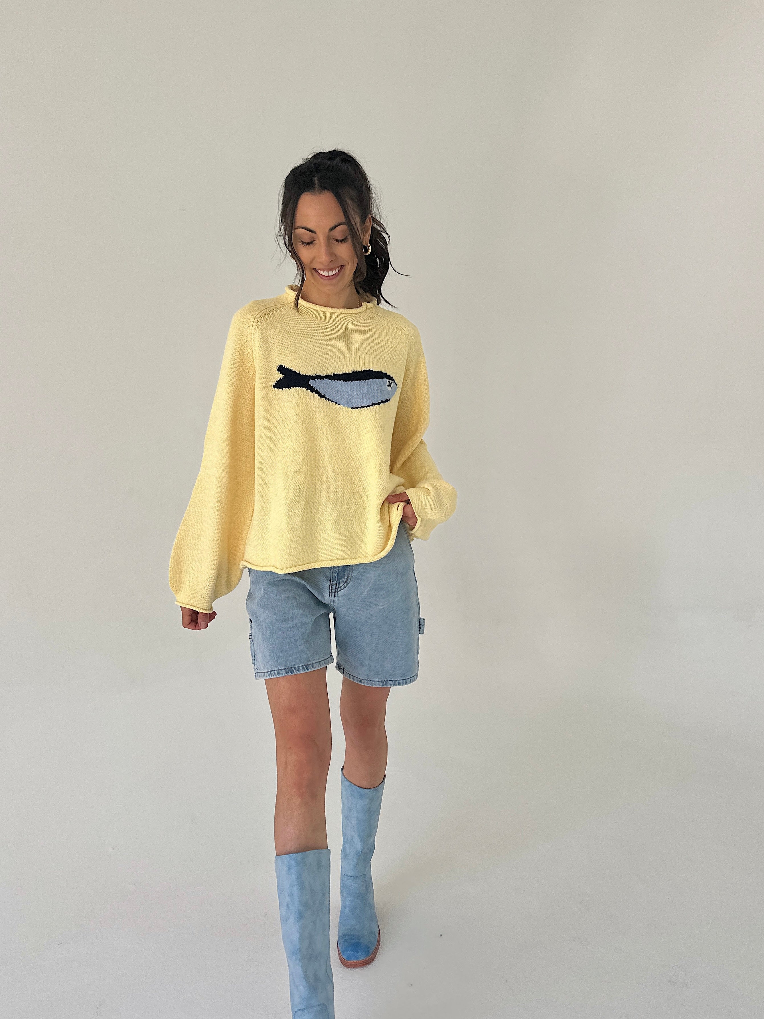Woman wearing a yellow sweater with a fish design, denim shorts, and blue boots on a plain background