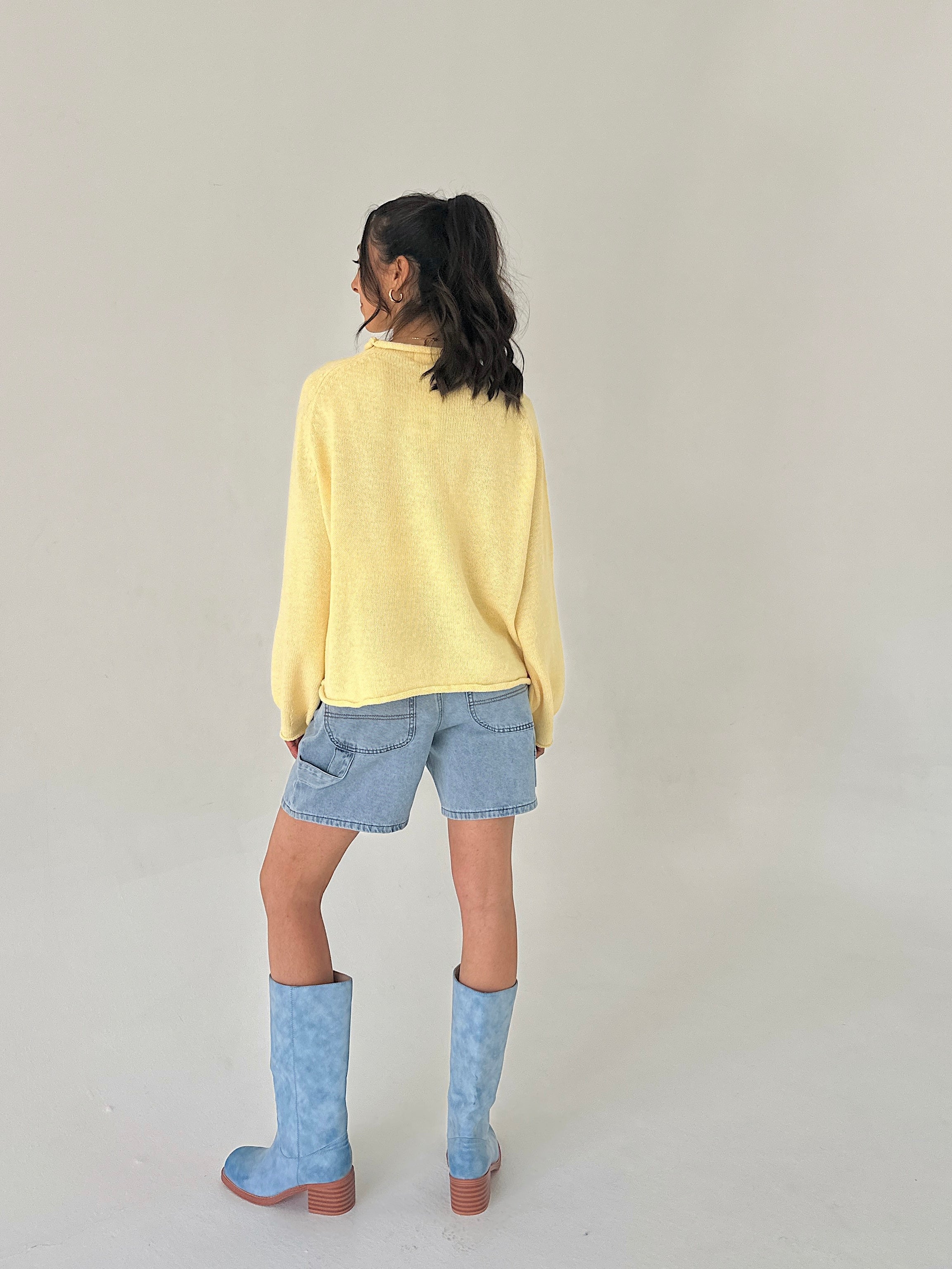 Person wearing a yellow sweater, denim shorts, and blue boots on a plain background from back
